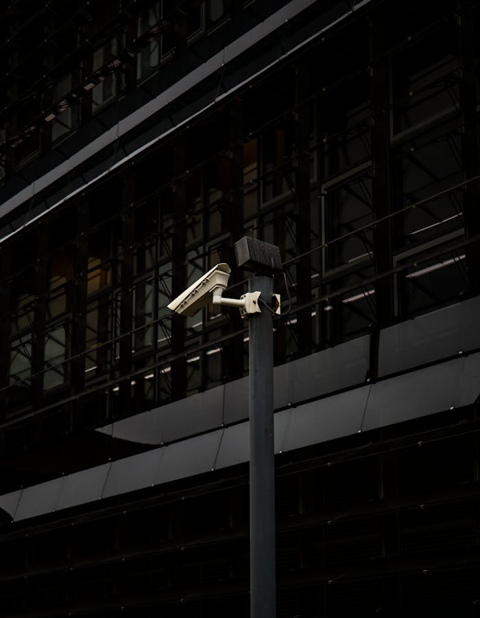 A surveillance camera mounted on a post against a modern urban building in a city setting.